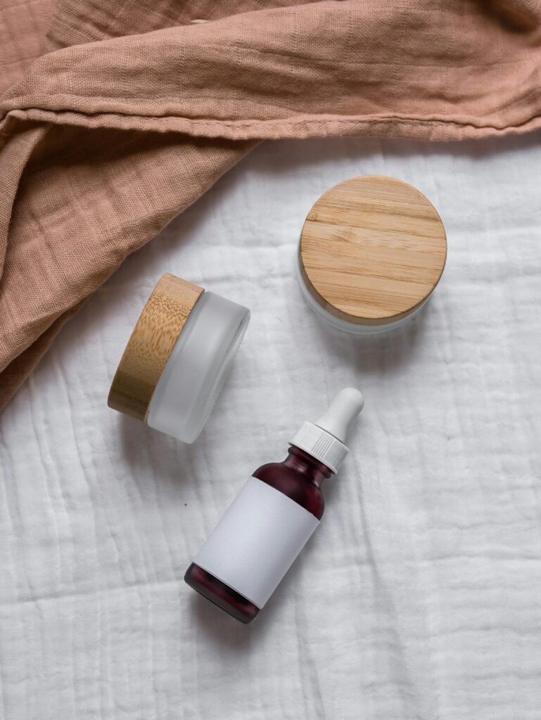 Flatlay of natural skincare jars and dropper bottle with wooden lids on a soft fabric background.