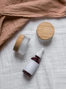 Flatlay of natural skincare jars and dropper bottle with wooden lids on a soft fabric background.